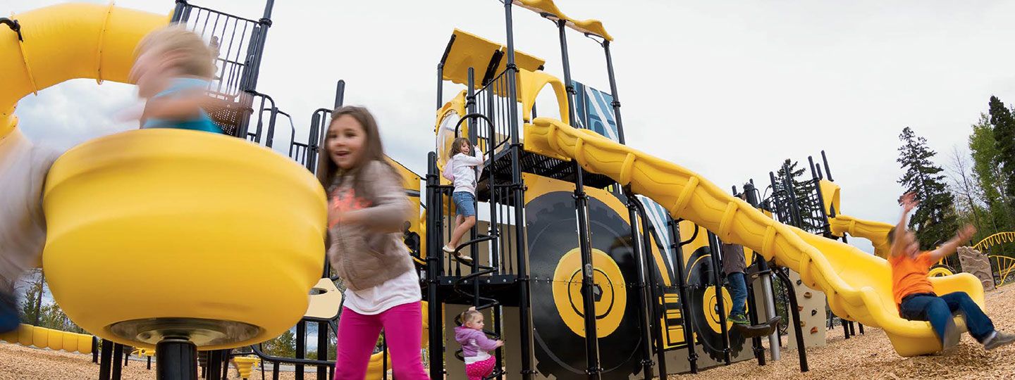 Children playing on yellow slides and climbing structures at a city park playground, featuring durable playground equipment for parks