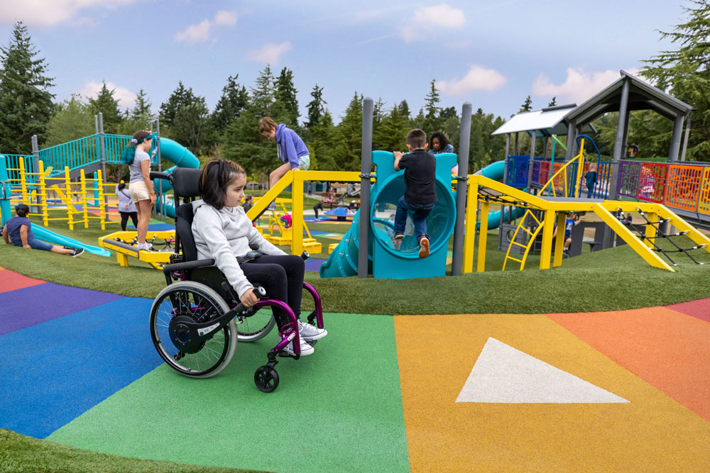 Young girl using a wheelchair navigating a colorful, wheelchair-accessible playground with ramps, slides, and inclusive equipment, while other children play nearby.
