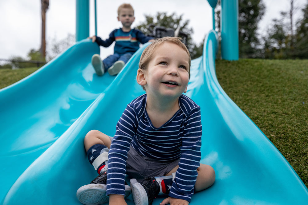 Young boy smiling at the bottom of an inclusive slide while another child follows behind, enjoying a play structure designed with accessible surfacing and supportive features for all abilities.