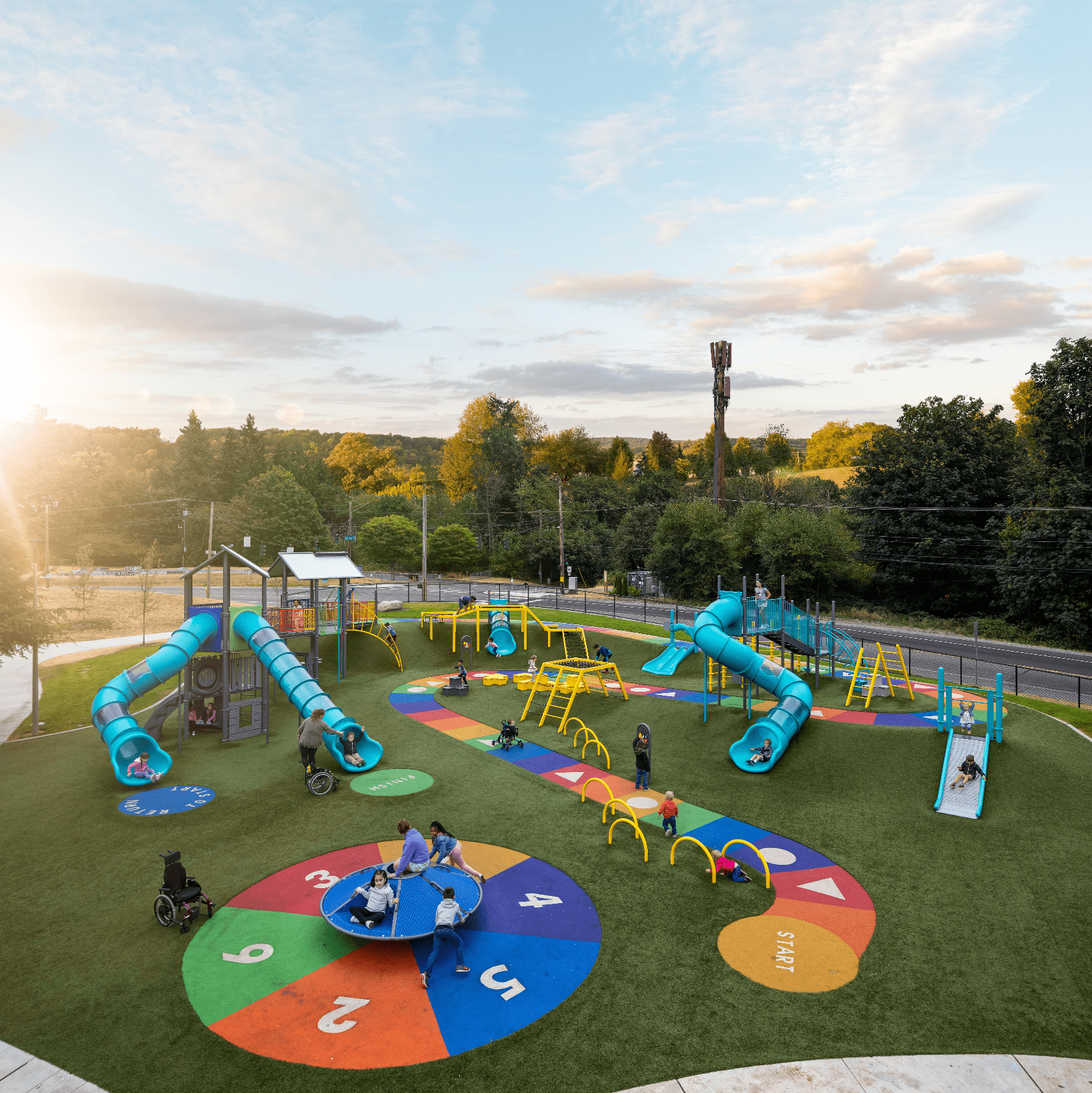 Wide view of an inclusive playground at sunset, featuring multiple tube slides, adaptive climbing structures, and children of all abilities—including wheelchair users—engaging in play on colorful, accessible surfacing.