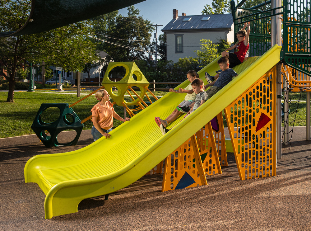 A four kids sliding down a Mighty Descent slide together while an adult watches from the side