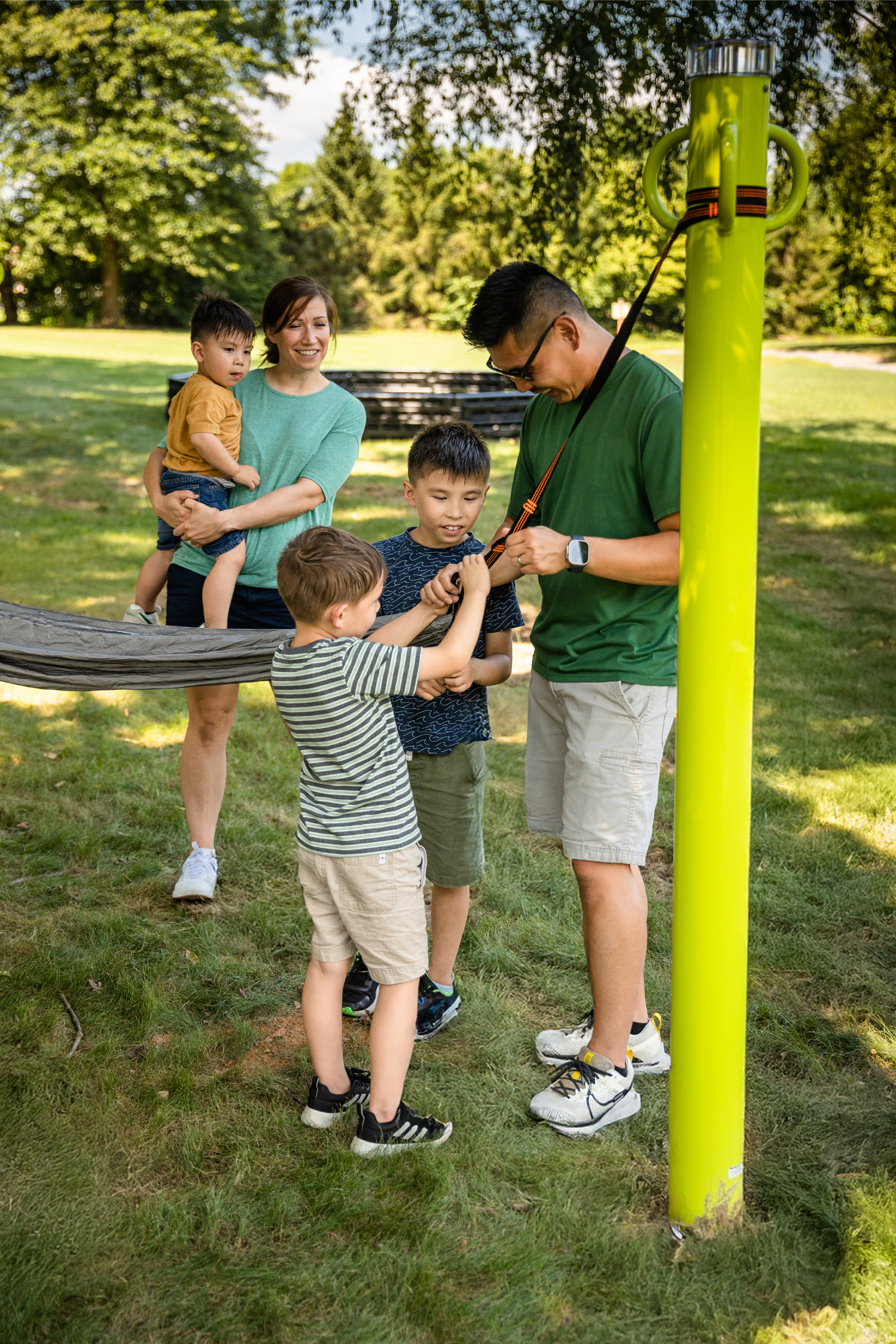 A dad and two sons attaching a hammock to a hammocking post while mom and another sibling watches